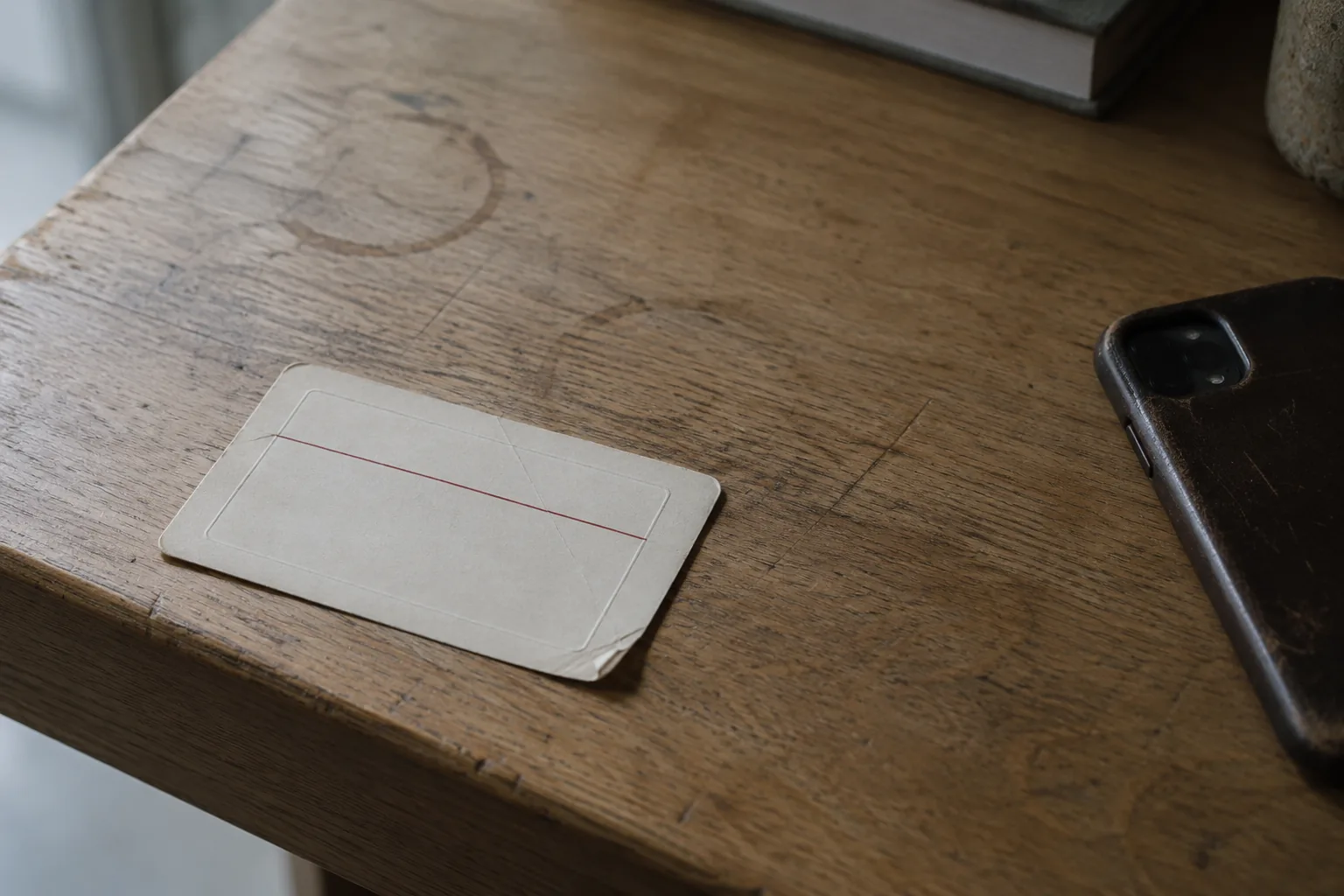 A plain cream library card lying on a scuffed oak table edge in low raking light. It carries a blind embossed panel and one thin red rule and nothing else. One corner is bent. The oak around it shows old ring marks and scratches, and a worn phone lies face down at the right edge of the frame.