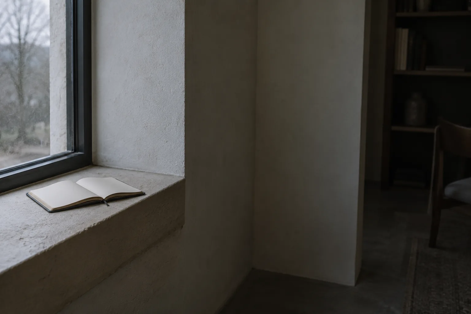 A blank open notebook resting on a deep plaster windowsill in the corner of a room, grey daylight at the window and the rest of the room falling away into shadow.