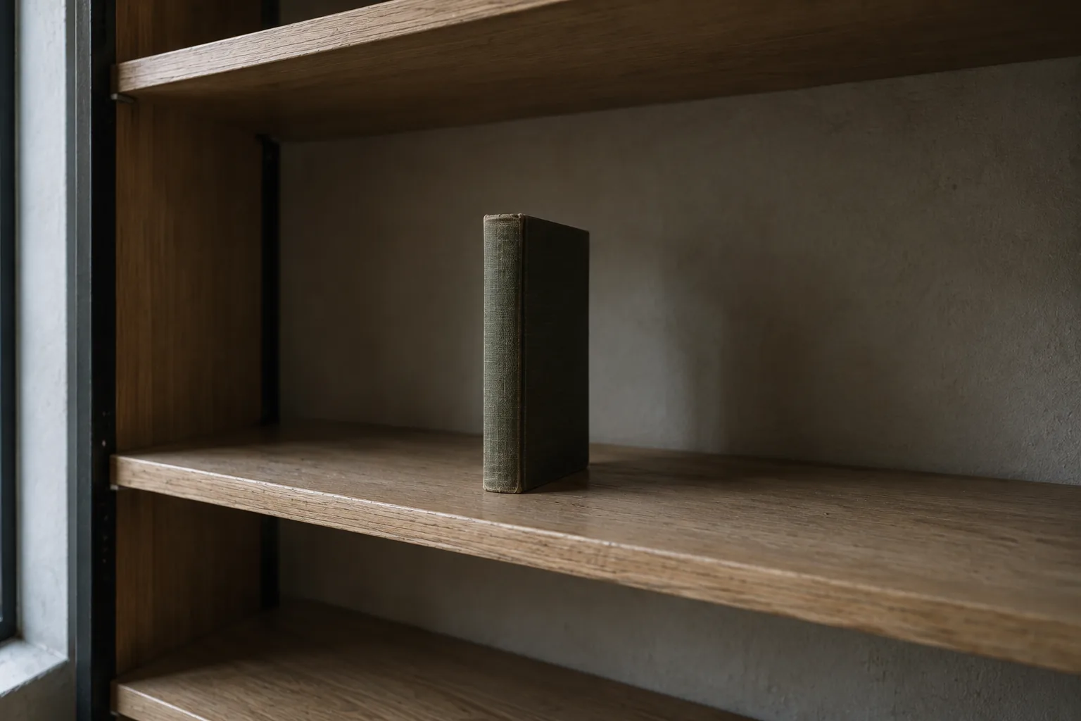 A single green cloth-bound book standing alone and upright in the middle of an otherwise completely empty pale oak shelf.